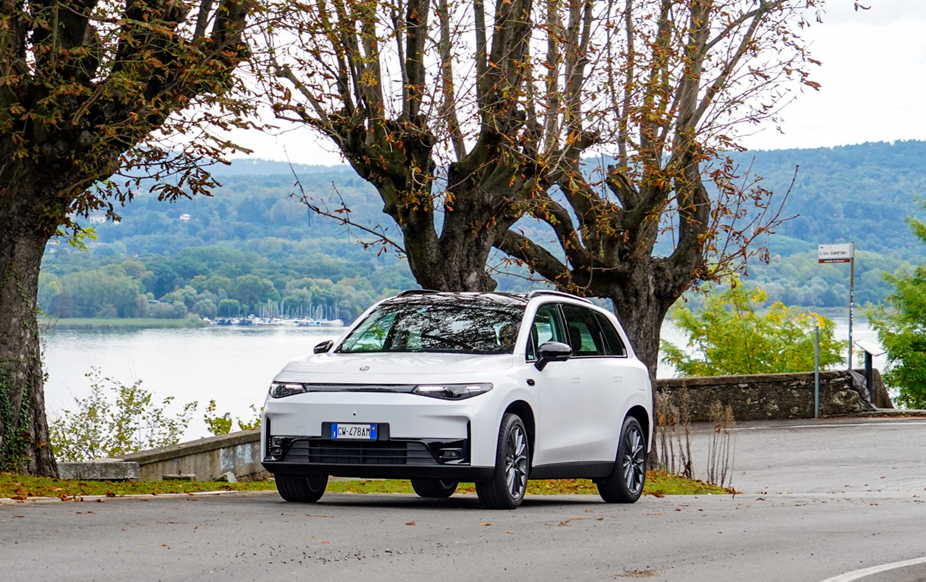 Ein weißes Auto steht auf einer Straße, die von Bäumen gesäumt ist. Im Hintergrund ist ein See mit einer bewaldeten Hügellandschaft sichtbar. Es ist ein bewölkter Tag.