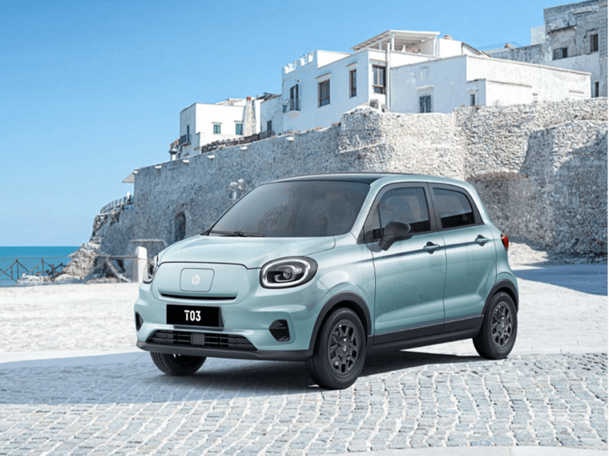 Ein hellblaues Auto steht auf einer gepflasterten Fläche vor einem Hintergrund aus weißen Gebäuden und einer Steinmauer, die an einen mediterranen Ort erinnert. Der Himmel ist klar und blau.